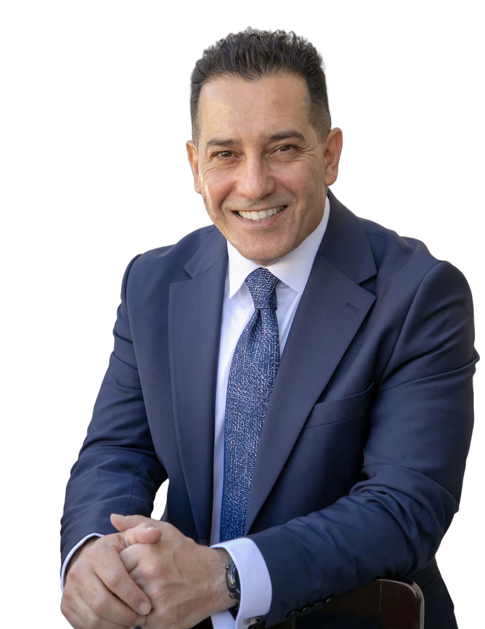 Cam Sourani, smiling, in a navy suit and blue tie, leaning forward with his hands clasped